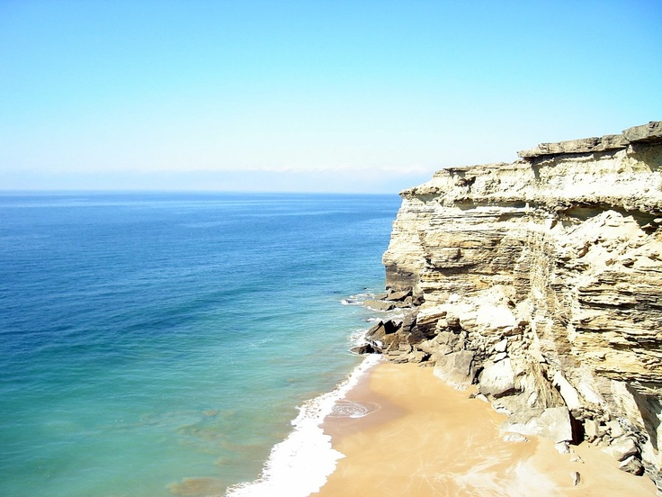 A scenic view of a coastline featuring sandy beach and steep limestone cliffs under a clear blue sky.