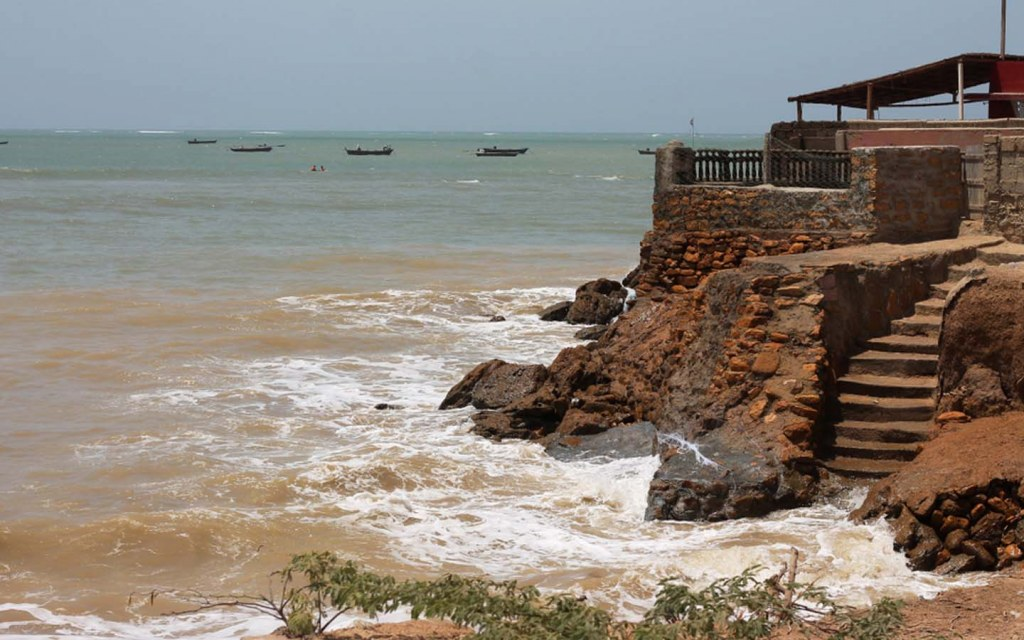A coastal scene showing water crashing against rocky shorelines, with some boats visible in the distance and a building structure partially overlooking the water.