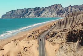 A scenic coastal road winding along a beach with cliffs and mountains in the background.