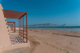 A beach scene under clear and blue sky with sandy shores, gentle waves, and mountains in the background, with open frames architectural structur near the beach.